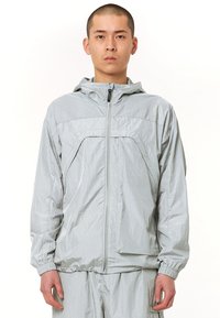 Jachetă ușoară, gri deschis, cu glugă, având un fermoar frontal complet și manșete elastice. Textură netedă, cu detalii de design minime.
