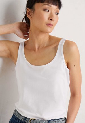 White sleeveless tank top made of soft fabric with a wide neckline, paired with a gray braided belt, and worn with blue denim jeans.
