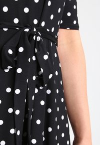 Czarna sukienka w białe kropki, z paskiem w talii i krótkimi rękawami. Tkanina wygląda na gładką i ma miękką teksturę.