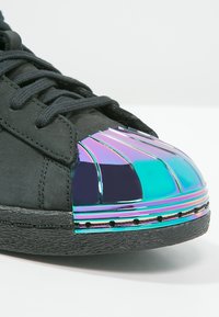 Black sneaker with a textured upper, featuring a metallic iridescent toe cap and contrasting black sole. Laces are dark and textured.