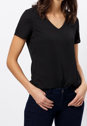 Kobieta w gładkiej, czarnej bluzce z krótkim rękawem i dekoltem w serek, włożonej do ciemnoniebieskich dżinsów, z rękami spoczywającymi blisko przednich kieszeni.