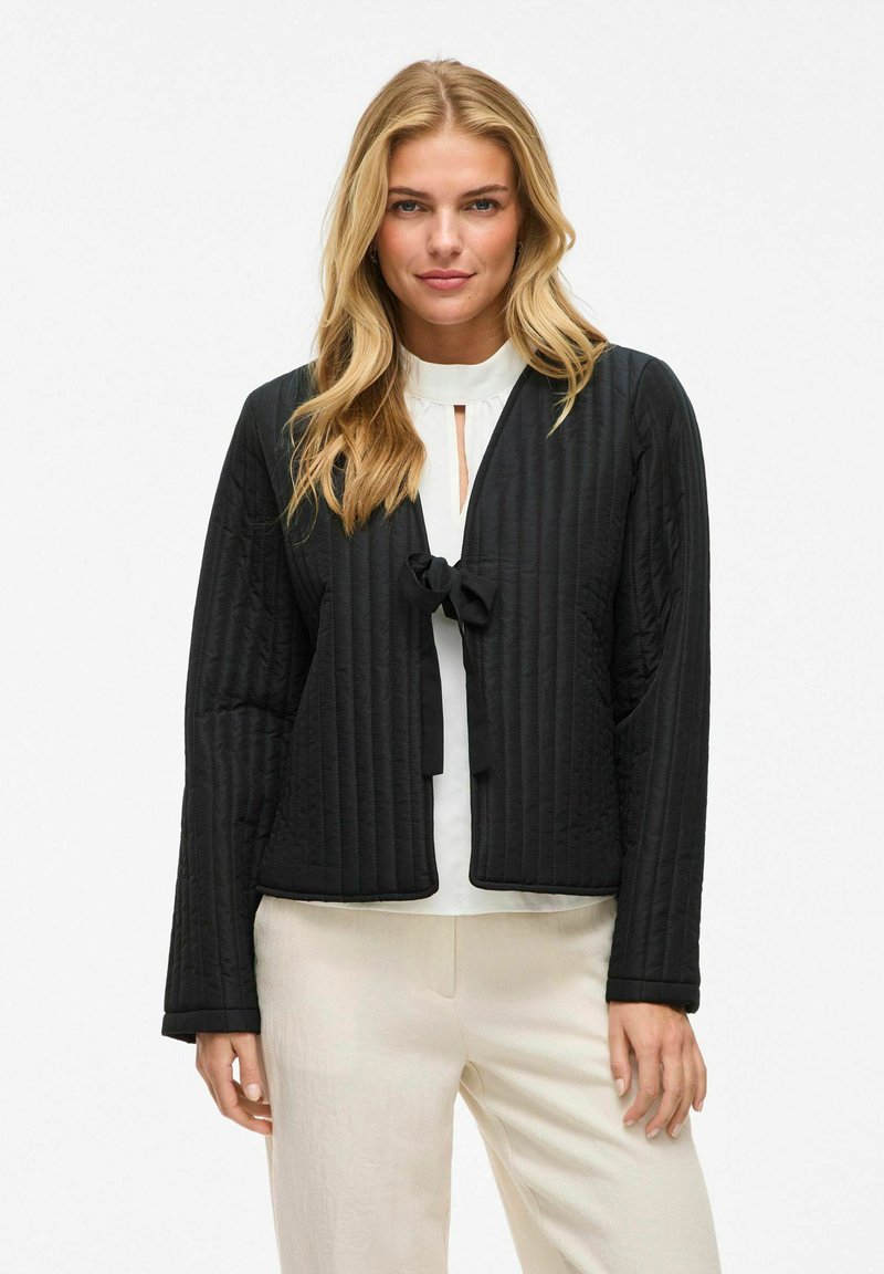 Svart quiltad jacka med långa ärmar och knytning framtill. Tyget har ett strukturerat mönster och skjortan bärs över en vit blus.