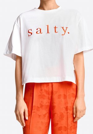 Witte cropped t-shirt met het woord "salty." in oranje letters. Losse pasvorm en korte mouwen, gecombineerd met oranje broek met patroon.