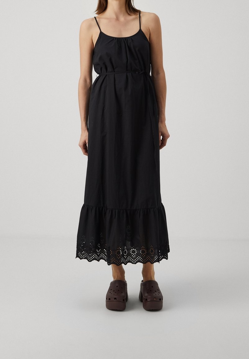 Femme portant une robe midi noire sans manches avec un ourlet en dentelle ajourée et des sabots marron à enfiler, debout devant un fond gris clair uni.