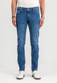 Mężczyzna w niebieskich jeansach typu slim-fit, białych trampkach, białej koszulce, srebrnym zegarku na lewej ręce oraz czarnej bransoletce na prawej ręce, stoi.