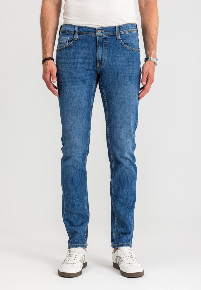 Mężczyzna w niebieskich jeansach typu slim-fit, białych trampkach, białej koszulce, srebrnym zegarku na lewej ręce oraz czarnej bransoletce na prawej ręce, stoi.