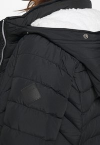 Svart vadderad jacka med quiltad design, huva fodrad med konstpäls och präglad logotyp på vänster axel. Har en silverfärgad dragkedja.