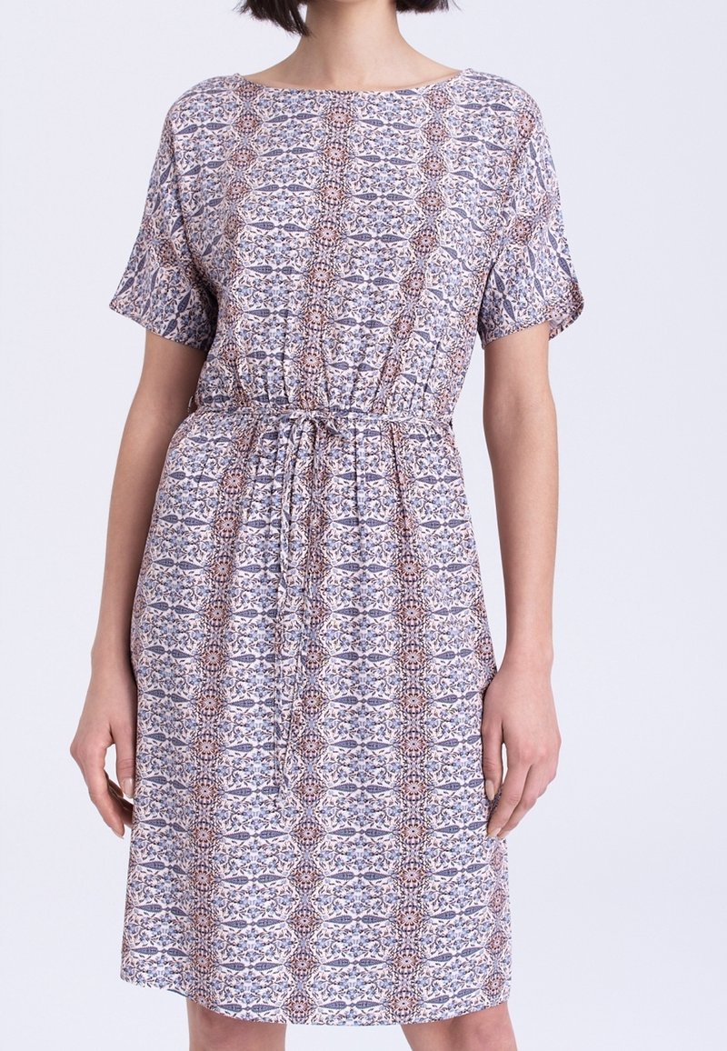 Femme portant une robe courte jusqu'aux genoux, à manches courtes, avec un motif géométrique répétitif et une taille à cordon.