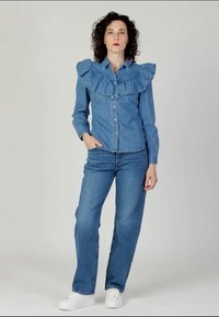 Denim shirt with large frilled collar, blue color, front buttons, worn with blue straight-leg jeans and white sneakers on a neutral background.
