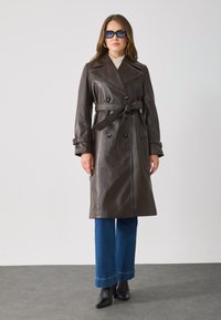Brown leather trench coat with double-breasted buttons, belted waist, wide lapels, and side pockets, paired with blue wide-leg jeans and black boots.