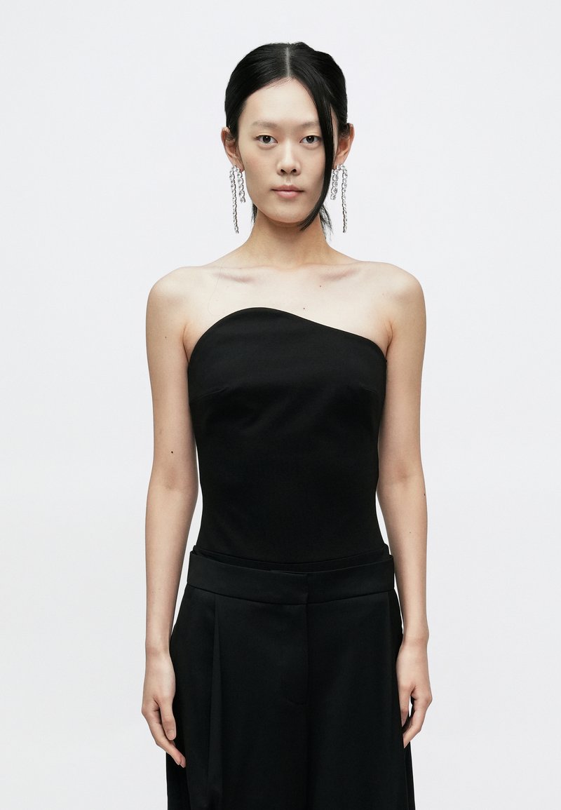 Melns strapless tops ar gludu tekstūru, ietilpīgu korsetveida augšdaļu un taisnu kakla izgriezumu. Kombinēts ar melnām platu kāju biksēm.