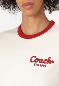 Creme-kleurig T-shirt met een rode ribgebreide kraag. Voorzien van geborduurde "Coach" en "New York" logo's in rood en bruin. De textuur is soepel.
