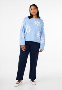 Hellblauer Strickpullover mit weißen Blumenmustern, lockere Passform und geripptem Saum, kombiniert mit dunkelblauen weit geschnittenen Jeans und weißen Ballerinas.