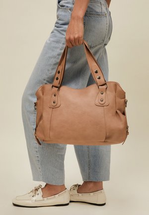 Person, die hellblaue Jeans und cremefarbene Loafers trägt, hält eine große hellbraune Lederhandtasche an den Griffen seitlich.