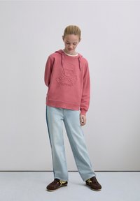 Pink hoodie with a skull and crossbones design, textured material, drawstring hood, paired with light blue jeans featuring side stripes.
