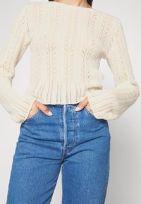 Strickpullover in Creme mit strukturiertem, offenem Muster und langen Ärmeln, kombiniert mit hochgeschnittenen, hellblauen Jeans.