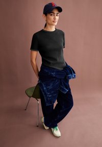 T-shirt noir à côtes, pantalons en velours côtelé bleu marine maintenus à la taille, et baskets beige avec des accents verts. L'arrière-plan est d'une teinte marron sourde.