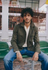 Khakijacke mit großen Taschen, über einem weißen Kurzarmoberteil getragen, kombiniert mit blauen Denim-Jeans. Auffällige Tattoos und Ringe an den Händen.