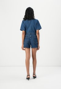 Macacão de ganga em azul escuro com mangas curtas, cintura ajustada e um corte descontraído. Apresenta dois bolsos traseiros e um design nas costas sem costuras.