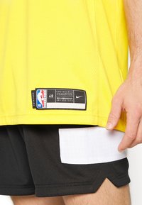 Žlutý basketbalový dres s síťovanou texturou, s černou etiketou s logem NBA a velikostí 48. Černé šortky s bílým mřížkovaným vzorem.