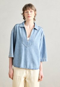 Top in denim azzurro con scollatura a V e maniche a metà. Design casual con dettagli di cucitura lungo i bordi. Abbinato a pantaloni di colore crema.