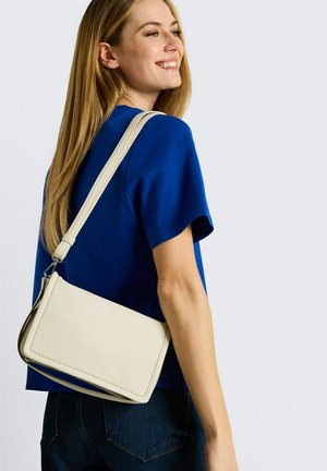 Vrouw in een blauw shirt met een witte schoudertas, glimlachend en zijwaarts kijkend tegen een effen achtergrond.