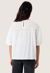 Weiße Bluse mit kurzen Ärmeln, strukturiertem Stoff, Schlüssel-ausschluss hinten und horizontalen Ausschnitten entlang der Schulter-nähte.