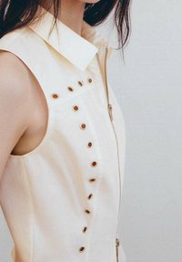 Beiges ärmelloses Kleid mit Kragen, ausgestattet mit einem Reißverschluss auf der Vorderseite und dekorativen Ösen entlang der linken Seite. Glatte Stofftextur.