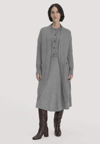Grauer gestrickter Cardigan mit offenem Front und zwei Taschen, über einem passenden Kleid getragen. Das Outfit zeichnet sich durch eine subtile Textur und eine lockere Passform aus.