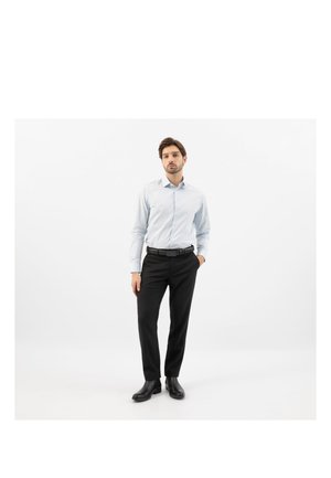 Uomo che indossa una camicia azzurro chiaro a maniche lunghe con bottoni, cintura nera, pantaloni neri e stivali neri, in piedi con una mano in tasca su sfondo bianco.