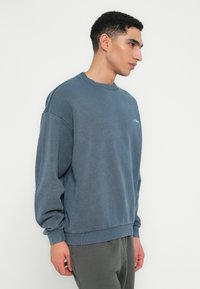 Sweater albastru cu guler rotund, umeri lăsați, manșete ribbate și tiv. Dispune de un logo subtil pe piept. Textură moale.