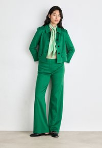 Green tailored suit with a cropped jacket and wide-leg trousers. Paired with a light green blouse and patterned scarf; features black shoes.