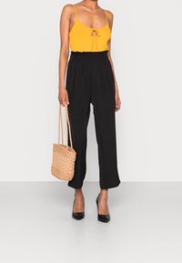 Svarta höga byxor med veck, kombinerat med en gul topp med spagettiband och en knut fram. Accessoarerna inkluderar en vävd beige handväska.