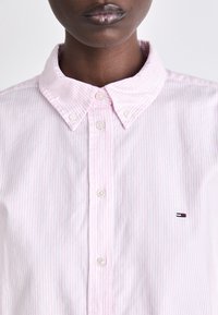 Camicia a righe rosa e bianche con colletto aperto, bottoni bianchi e una piccola toppa con logo sul petto.
