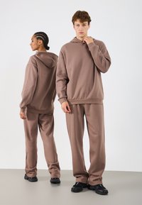 Brązowa bluza z kapturem i dopasowane spodnie dresowe wykonane z miękkiej, teksturowanej tkaniny. Luźny krój z prążkowanymi mankietami i pasem. Czarne buty.