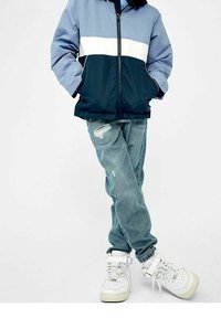 Blå och marinblå färgblockerad jacka med dragkedja, i kombination med ljusblå slitna jeans och vita hög-top sneakers.