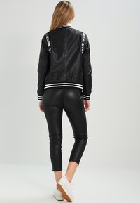 Kvinna iklädd en svart läderbomberjacka, svarta tighta byxor och vita sneakers, sedd bakifrån mot en ljus bakgrund.