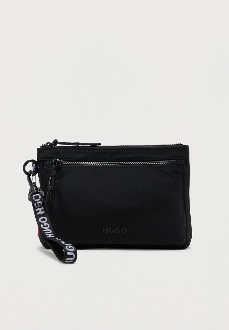 Μαύρο τσαντάκι Hugo με μπροστινή τσέπη με φερμουάρ και αποσπώμενο λουράκι καρπού με λογότυπο, σε απλό ανοιχτόχρωμο φόντο.