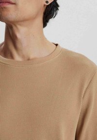 Beige bársonyos kötött kerek nyakú pulóver, textúrált felülettel, széles nyakkal és enyhe rugalmassággal. Fekete szegecses fülbevaló látható.