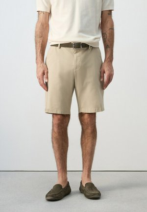 Mann trägt beige knielange Shorts, olivgrünen Gürtel, cremefarbenes T-Shirt und olivgrüne Loafers, steht vor einem schlichten Hintergrund.
