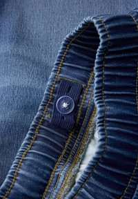 Denimska tkanina tamnoplave nijanse, s nabranim strukom, kontrastnim žutim šavovima i prozirnim dugmetom za zatvaranje.