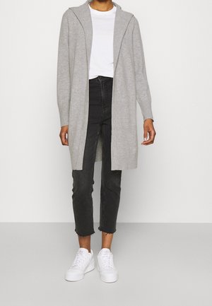 Personne portant un long cardigan gris par-dessus une chemise blanche, un jean noir raccourci et des baskets blanches, debout devant un fond uni.