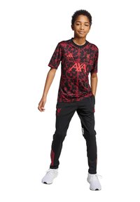 Camisa deportiva del Liverpool FC en rojo y negro con un patrón floral, combinada con pantalones negros con rayas rojas y un logo. Zapatillas deportivas blancas.