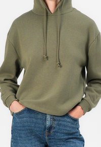 Sweat à capuche vert olive avec cordons, coupe ample, manches longues et poignets côtelés, associé à un jean bleu. Texture lisse.