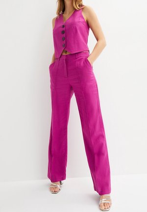 Femme portant un gilet sans manches rose vif à boutons et un pantalon large assorti, talons blancs à brides, mains dans les poches, debout contre un mur blanc.