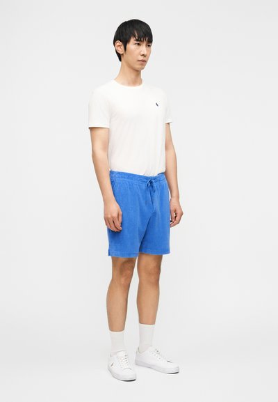 Άνδρας όρθιος που φοράει άσπρο κοντομάνικο μπ λουζάκι, μπλε σορτς με κορδόνι, άσπρες κάλτσες και άσπρα αθλητικά παπούτσια, μπροστά σε απλό φόντο.