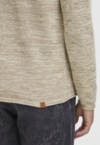 Beige marled knit sweater worn with black washed denim jeans, showing the lower back and right arm of the person.