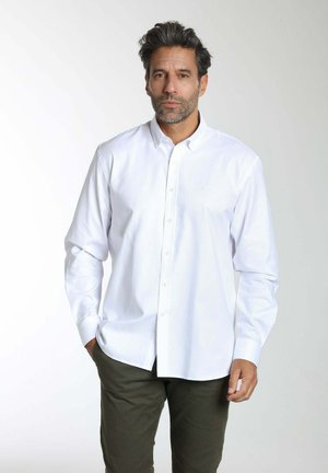 Homme aux cheveux foncés et à la barbe, portant une chemise blanche boutonnée et un pantalon vert foncé, debout devant un fond clair uni.