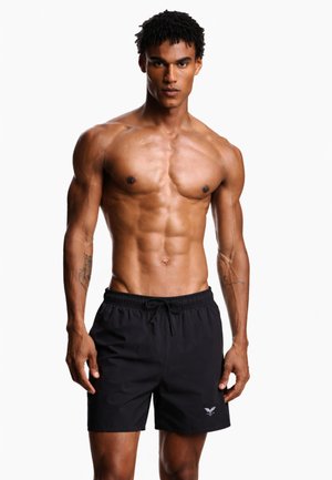 Αθλητικός άνδρας με τατουάζ που φοράει μαύρο μαγιό, στέκεται μπροστά σε μια απλή λευκή επιφάνεια με ουδέτερη έκφραση.