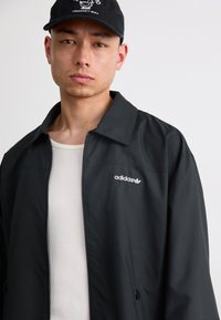 Black zip-up jacket with a collared design, featuring a white Adidas logo on the left chest. Worn over a light ribbed shirt.